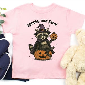 Peut inclure: T-shirt rose clair pour enfant avec un motif d'Halloween. Le motif repr&eacute;sente un raton laveur portant un chapeau et une cape de sorci&egrave;re, assis sur une citrouille sculpt&eacute;e et tenant une lanterne. Le texte au-dessus dit "Spooky and Feral."