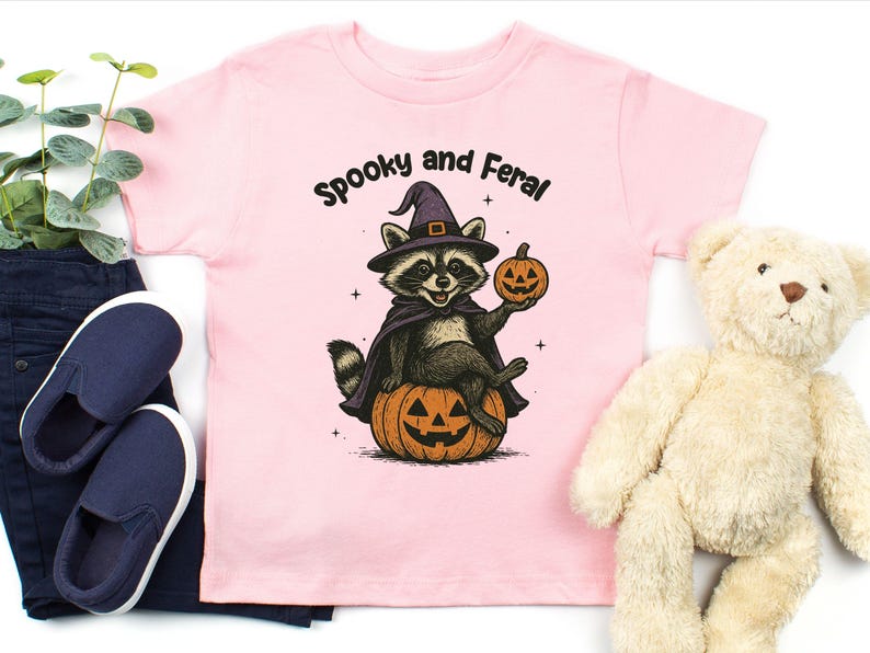 Peut inclure: T-shirt rose clair pour enfant avec un motif d'Halloween. Le motif repr&eacute;sente un raton laveur portant un chapeau et une cape de sorci&egrave;re, assis sur une citrouille sculpt&eacute;e et tenant une lanterne. Le texte au-dessus dit "Spooky and Feral."