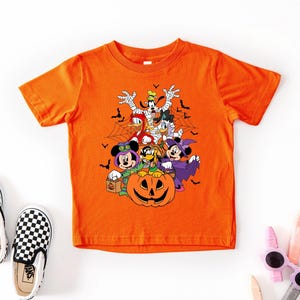 Op de afbeelding: Oranje T-shirt met korte mouwen met een Halloween-thema met stripfiguren in kostuums, waaronder een mummie, heks en vampier. Een pompoen staat in het midden. Zwart-wit geblokte instappers aan de linkerkant.