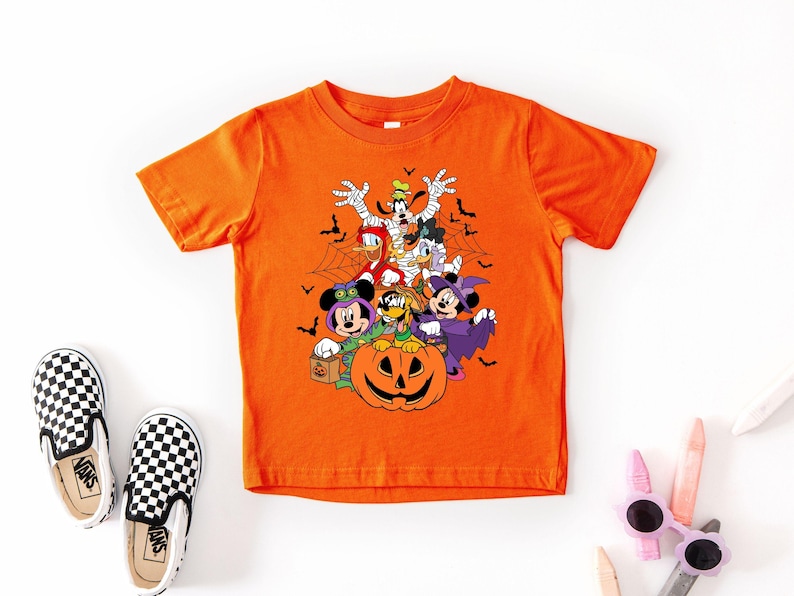 Op de afbeelding: Oranje T-shirt met korte mouwen met een Halloween-thema met stripfiguren in kostuums, waaronder een mummie, heks en vampier. Een pompoen staat in het midden. Zwart-wit geblokte instappers aan de linkerkant.