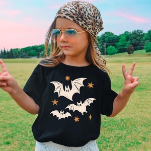 May include: A black short-sleeved t-shirt featuring a graphic print of three cream-colored bats and several small orange stars. The shirt has a crew neckline and a straight hem, providing a simple and classic fit for casual daily wear.