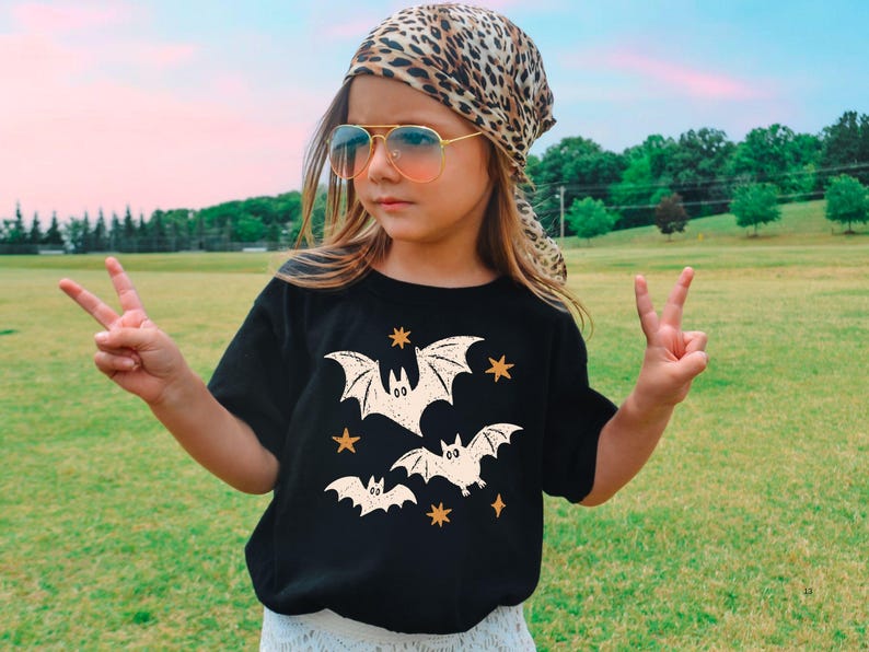 May include: A black short-sleeved t-shirt featuring a graphic print of three cream-colored bats and several small orange stars. The shirt has a crew neckline and a straight hem, providing a simple and classic fit for casual daily wear.