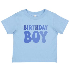 May include: A pale blue t-shirt featuring the text "BIRTHDAY BOY" in varying shades of blue. The shirt has a crew neck and short sleeves, ideal for a child's birthday party.