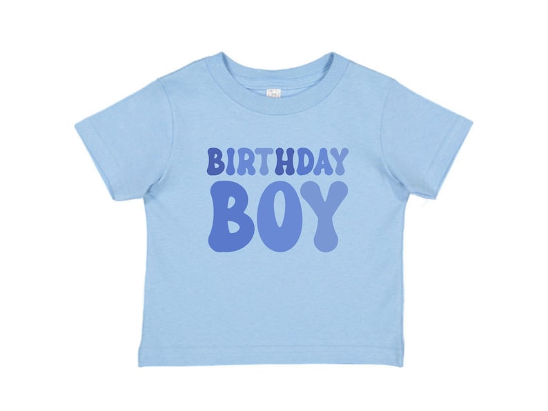 May include: A pale blue t-shirt featuring the text "BIRTHDAY BOY" in varying shades of blue. The shirt has a crew neck and short sleeves, ideal for a child's birthday party.