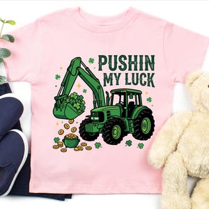 May include: A light pink t-shirt featuring a green tractor with a bucket full of shamrocks and gold coins. The text "PUSHIN MY LUCK" is printed above the tractor. The shirt is paired with navy blue shoes and a teddy bear.