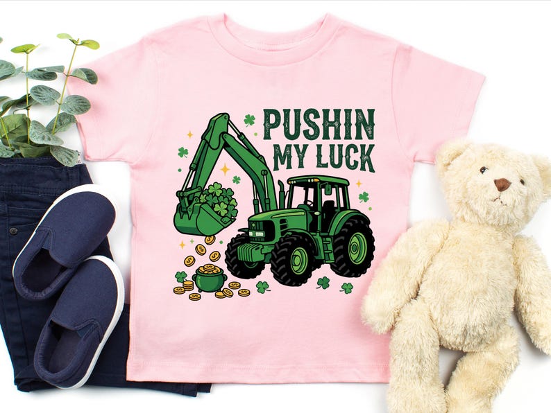 May include: A light pink t-shirt featuring a green tractor with a bucket full of shamrocks and gold coins. The text "PUSHIN MY LUCK" is printed above the tractor. The shirt is paired with navy blue shoes and a teddy bear.