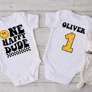 May include: Two white baby onesies. One has the text "ONE HAPPY DUDE" with a smiley face and checkered pattern. The other has the name "OLIVER" and a yellow number 1 with smiley faces. A stuffed bunny is next to the onesie.