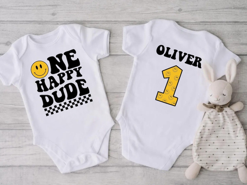May include: Two white baby onesies. One has the text "ONE HAPPY DUDE" with a smiley face and checkered pattern. The other has the name "OLIVER" and a yellow number 1 with smiley faces. A stuffed bunny is next to the onesie.