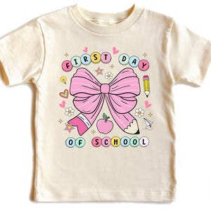 May include: A cream-coloured t-shirt with a large pink bow, apple, and pencil design. The words "FIRST DAY OF SCHOOL" are written in a colourful, circular design. Additional decorative elements include hearts, stars, and flowers.