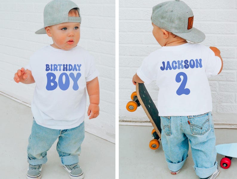 May include: A young child in a white t-shirt with the words "BIRTHDAY BOY" in blue. The child wears a grey cap, blue jeans, and trainers. The back of the shirt says "JACKSON 2". The child is holding a skateboard.
