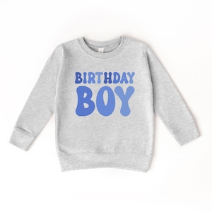 May include: A grey sweatshirt with long sleeves. The front displays the words "BIRTHDAY BOY" in blue, retro lettering. This children's sweatshirt is designed for a birthday celebration. The design is simple and eye-catching.
