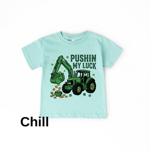 May include: A light blue t-shirt featuring a green tractor with a bucket full of shamrocks and gold coins. The text "Pushin' My Luck" is above the tractor. The word "Chill" is at the bottom.