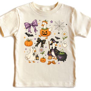 May include: A cream-coloured short-sleeve t-shirt featuring a colourful Halloween-themed graphic print. The design includes illustrations of ducks dressed in costumes, pumpkins, a spider web, bats, bows, a ghost, and a bubbling cauldron.