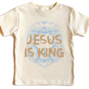 May include: Cream-colored t-shirt featuring a lion's head with a blue mane and the text "JESUS IS KING" in a tan color. The shirt is a short-sleeved crew neck.