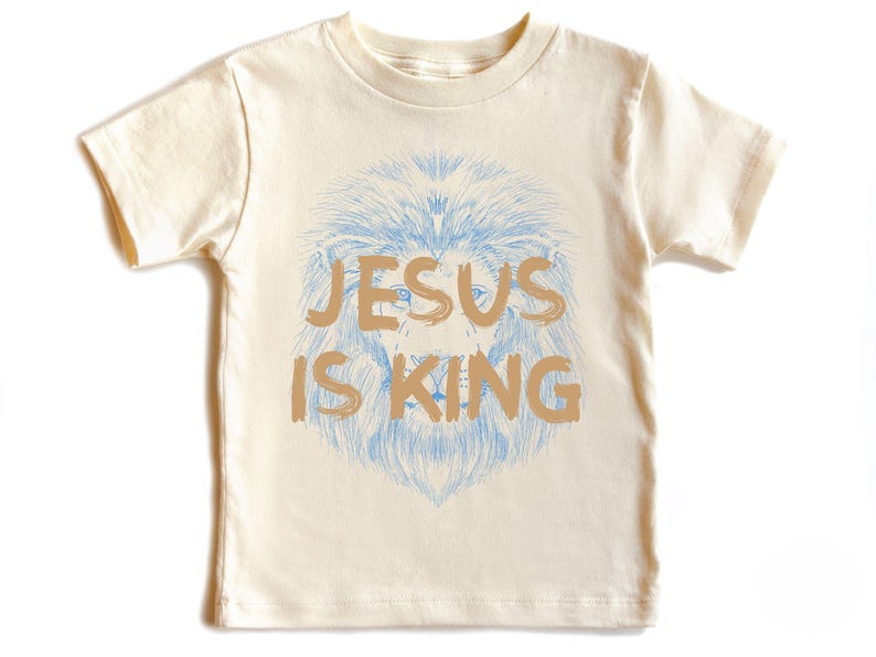 May include: Cream-colored t-shirt featuring a lion's head with a blue mane and the text "JESUS IS KING" in a tan color. The shirt is a short-sleeved crew neck.