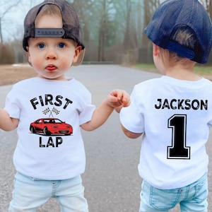 Peut inclure: Deux jeunes enfants portant des t-shirts blancs et des casquettes de baseball. L'un des t-shirts affiche "FIRST LAP" avec une voiture rouge. L'autre t-shirt porte "JACKSON" et le chiffre "1" au dos. Ils portent des jeans bleus.