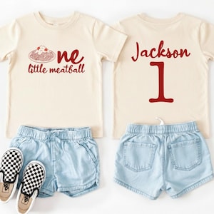 May include: A cream-colored toddler outfit featuring a t-shirt with a red graphic of spaghetti and the words "one little meatball." The back of the shirt has the name "Jackson" and the number "1" in red. Includes denim shorts and checkered sneakers.