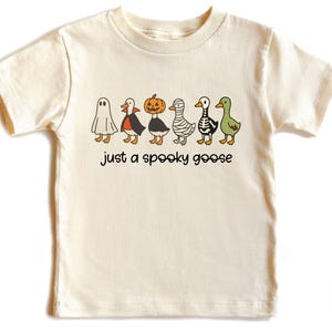 May include: Cream-colored t-shirt featuring a Halloween-themed graphic. The design includes six cartoon geese dressed as a ghost, vampire, pumpkin, mummy, skeleton, and green goose. The text "just a spooky goose" is below the geese.