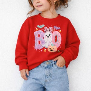 Puede incluir: Una sudadera roja de manga larga con un estampado gr&aacute;fico de la palabra BOO en letras rosas grandes. El dise&ntilde;o incluye un fantasma blanco con un lazo rosa de lunares, una calabaza naranja sonriente, peque&ntilde;os murci&eacute;lagos y caramelos.