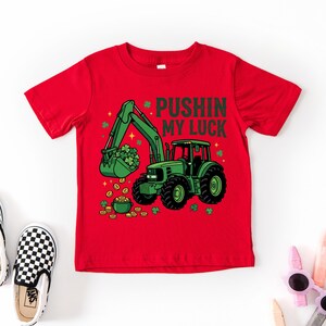 May include: Red t-shirt featuring a green tractor with a bucket full of shamrocks and a pot of gold. The text "PUSHIN MY LUCK" is above the tractor. The shirt is paired with black and white checkered slip-on shoes.