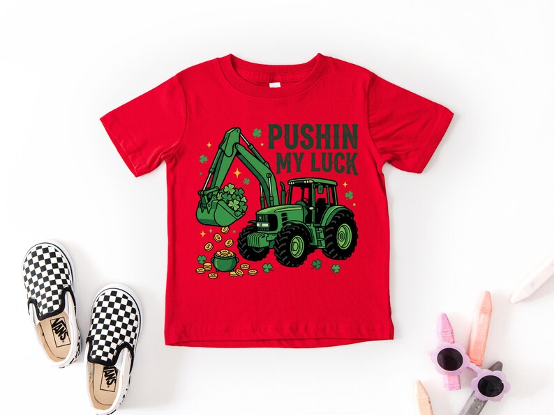 May include: Red t-shirt featuring a green tractor with a bucket full of shamrocks and a pot of gold. The text "PUSHIN MY LUCK" is above the tractor. The shirt is paired with black and white checkered slip-on shoes.
