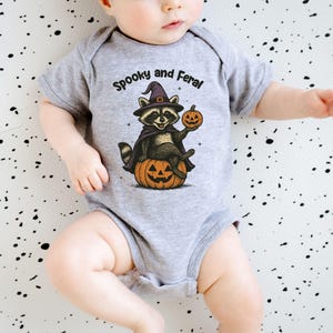 Peut inclure: Un body de b&eacute;b&eacute; gris avec le texte "Spooky and Feral" au-dessus d'un raton laveur de dessin anim&eacute; portant un chapeau de sorci&egrave;re et une cape, tenant une citrouille et assis sur une citrouille. Le fond est blanc avec des &eacute;claboussures noires.