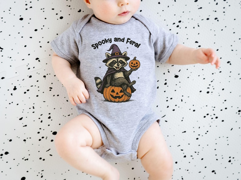 Peut inclure: Un body de b&eacute;b&eacute; gris avec le texte "Spooky and Feral" au-dessus d'un raton laveur de dessin anim&eacute; portant un chapeau de sorci&egrave;re et une cape, tenant une citrouille et assis sur une citrouille. Le fond est blanc avec des &eacute;claboussures noires.