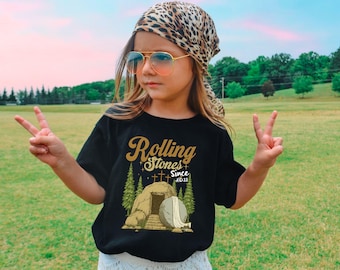 May include: Black t-shirt with the text "Rolling Stones Since AD 33" in gold lettering. The shirt features a graphic of a tomb with a stone and crosses. The person is wearing sunglasses and a leopard print scarf.