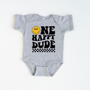 May include: A gray baby onesie with the text "ONE HAPPY DUDE" in black, featuring a yellow smiley face and a black and white checkered pattern. The onesie is short-sleeved.