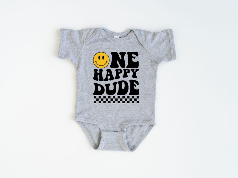 May include: A gray baby onesie with the text "ONE HAPPY DUDE" in black, featuring a yellow smiley face and a black and white checkered pattern. The onesie is short-sleeved.