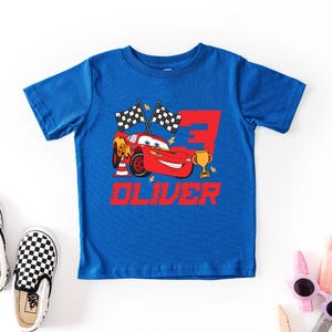 May include: A blue t-shirt featuring a red race car graphic with the number 3, checkered flags, and the name "OLIVER" in red. Black and white checkered slip-on shoes are next to the shirt.