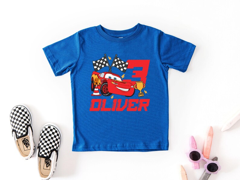 May include: A blue t-shirt featuring a red race car graphic with the number 3, checkered flags, and the name "OLIVER" in red. Black and white checkered slip-on shoes are next to the shirt.