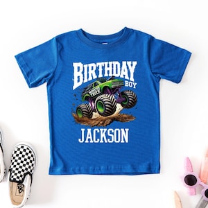 Pode incluir: Uma camiseta azul royal com um gr&aacute;fico de um monster truck verde e roxo. O texto na camisa diz "BIRTHDAY BOY" acima de "MONSTER TRUCK" e o nome "JACKSON". Sapatos slip-on xadrez est&atilde;o &agrave; esquerda.