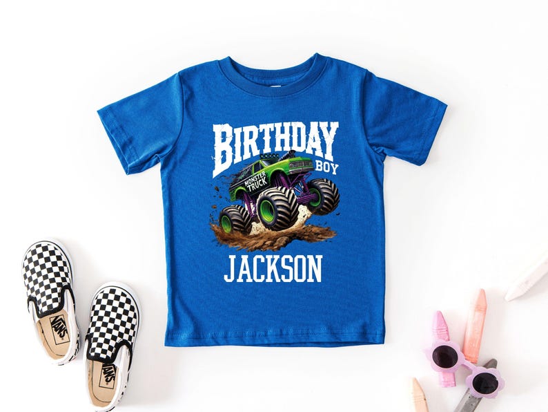 Pode incluir: Uma camiseta azul royal com um gr&aacute;fico de um monster truck verde e roxo. O texto na camisa diz "BIRTHDAY BOY" acima de "MONSTER TRUCK" e o nome "JACKSON". Sapatos slip-on xadrez est&atilde;o &agrave; esquerda.