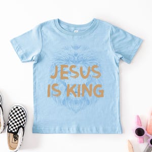 May include: Light blue short-sleeved t-shirt with the text "JESUS IS KING" in gold. The shirt features a lion's head design. Checkered black and white slip-on shoes are next to the shirt.