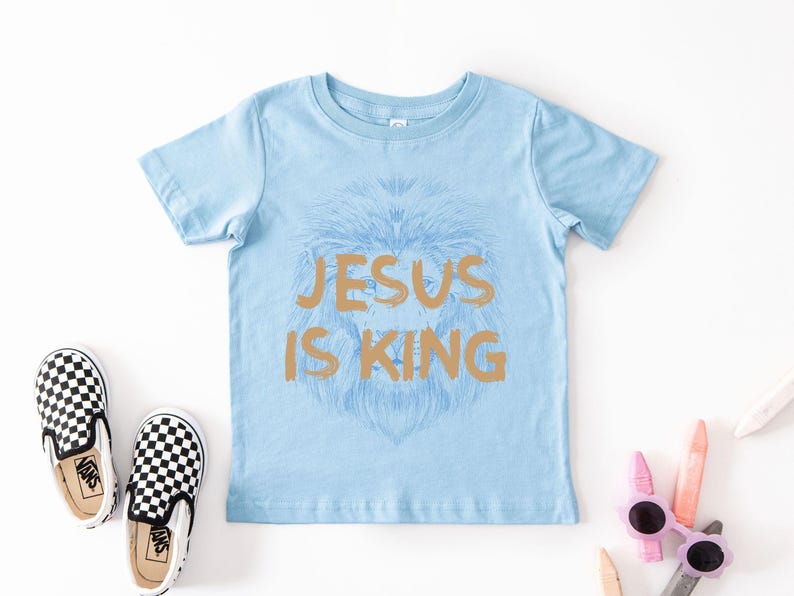 May include: Light blue short-sleeved t-shirt with the text "JESUS IS KING" in gold. The shirt features a lion's head design. Checkered black and white slip-on shoes are next to the shirt.