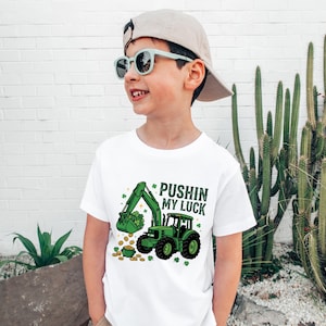 May include: White t-shirt with a green excavator graphic and the text "Pushin' my Luck". The excavator is scooping up gold coins and shamrocks. The shirt is worn by a child wearing sunglasses and a cap.