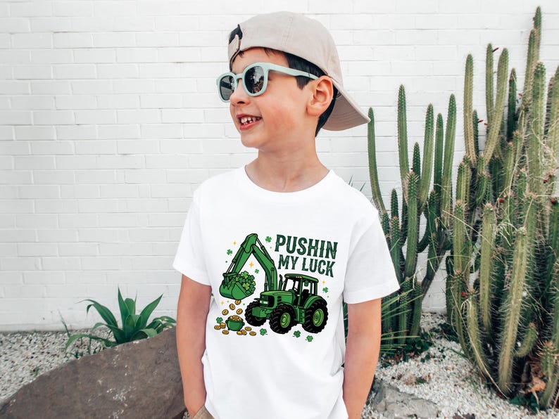 May include: White t-shirt with a green excavator graphic and the text "Pushin' my Luck". The excavator is scooping up gold coins and shamrocks. The shirt is worn by a child wearing sunglasses and a cap.