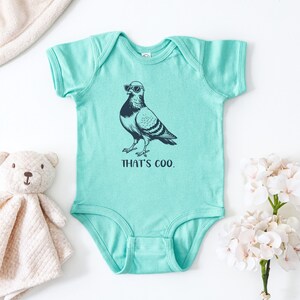 May include: A turquoise baby onesie with a graphic of a pigeon wearing sunglasses. The text "THAT'S COO." is printed below the bird. A stuffed teddy bear and a white blanket are in the background.