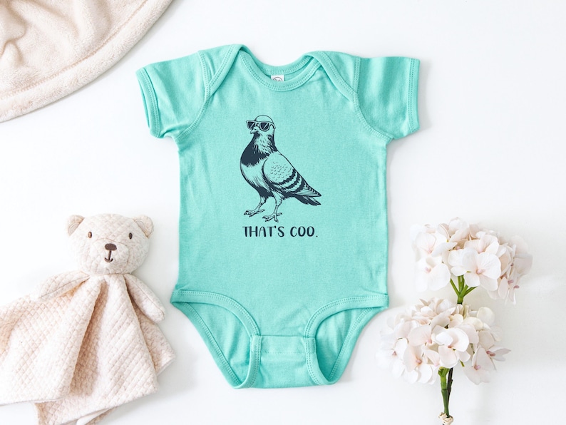 May include: A turquoise baby onesie with a graphic of a pigeon wearing sunglasses. The text "THAT'S COO." is printed below the bird. A stuffed teddy bear and a white blanket are in the background.