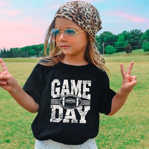 K&ouml;nnte beinhalten: Ein schwarzes T-Shirt mit den Worten "GAME DAY" und einer Football-Grafik in Wei&szlig;. Die Person tr&auml;gt ein Leopardenmuster-Tuch, eine Sonnenbrille und zeigt mit den Fingern ein Friedenszeichen.