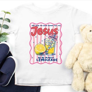 May include: White t-shirt with a colorful graphic featuring the word "Jesus" in pink, a glass of lemonade, and a lemon slice. The text "life gave me some lemons but my Jesus HE BE MAKIN' LEMONADE" is also included. Navy blue slip-on shoes and a teddy bear are also in the image.