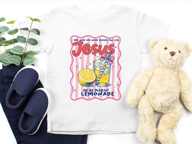 May include: White t-shirt with a colorful graphic featuring the word "Jesus" in pink, a glass of lemonade, and a lemon slice. The text "life gave me some lemons but my Jesus HE BE MAKIN' LEMONADE" is also included. Navy blue slip-on shoes and a teddy bear are also in the image.