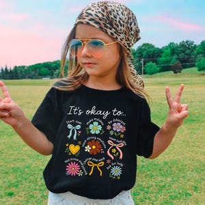 May include: A black t-shirt with the text "It's okay to..." and various floral and bow graphics. The shirt is worn by a person wearing sunglasses and a leopard print scarf. The person is making a peace sign with their fingers.