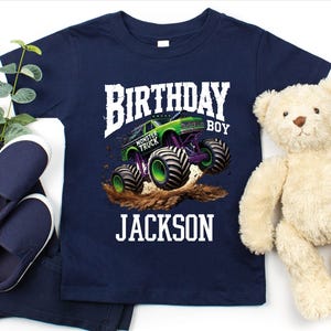 Pode incluir: Camiseta azul marinho com um gr&aacute;fico de caminh&atilde;o monstro colorido. O design inclui o texto "BIRTHDAY BOY" e o nome "JACKSON". A imagem inclui sapatos e cal&ccedil;as azul marinho e um ursinho de pel&uacute;cia.