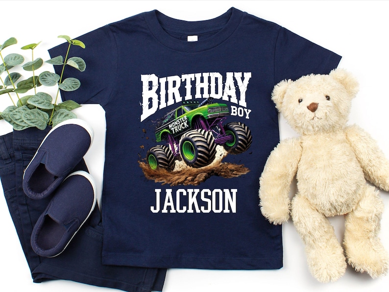 Pode incluir: Camiseta azul marinho com um gr&aacute;fico de caminh&atilde;o monstro colorido. O design inclui o texto "BIRTHDAY BOY" e o nome "JACKSON". A imagem inclui sapatos e cal&ccedil;as azul marinho e um ursinho de pel&uacute;cia.