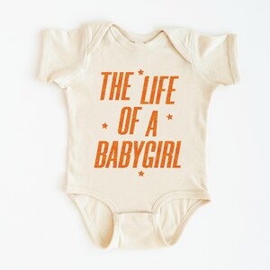 Pode incluir: Body de beb&eacute; creme com o texto laranja "THE LIFE OF A BABYGIRL" e estrelas. A pe&ccedil;a &uacute;nica &eacute; projetada para beb&ecirc;s e crian&ccedil;as pequenas.