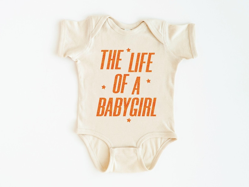 Pode incluir: Body de beb&eacute; creme com o texto laranja "THE LIFE OF A BABYGIRL" e estrelas. A pe&ccedil;a &uacute;nica &eacute; projetada para beb&ecirc;s e crian&ccedil;as pequenas.