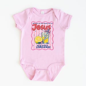 May include: A light pink baby bodysuit with a graphic of a lemonade drink, a lemon slice, and the text "Jesus He Be Makin' Lemonade". The design is in a pink and red frame.