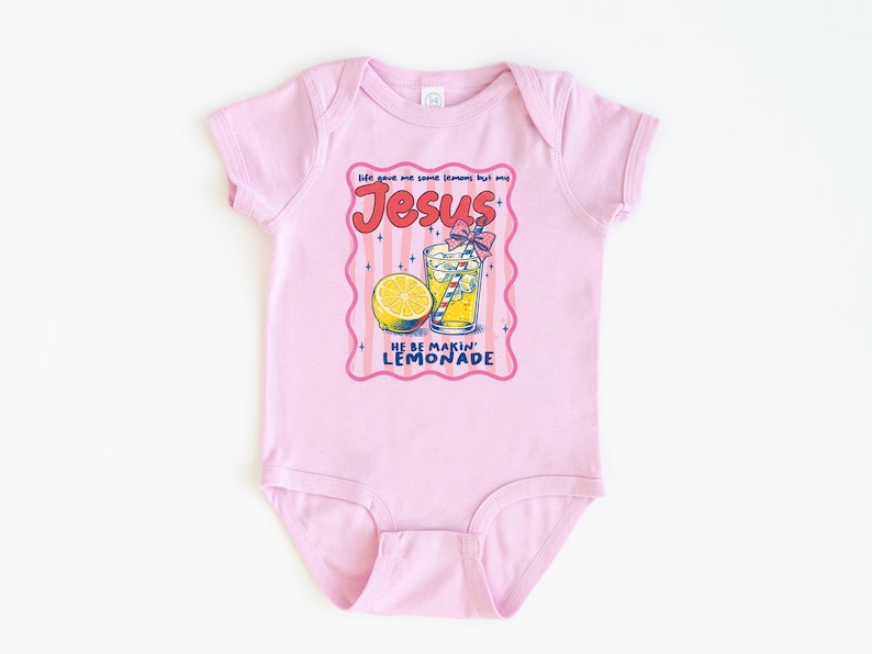 May include: A light pink baby bodysuit with a graphic of a lemonade drink, a lemon slice, and the text "Jesus He Be Makin' Lemonade". The design is in a pink and red frame.